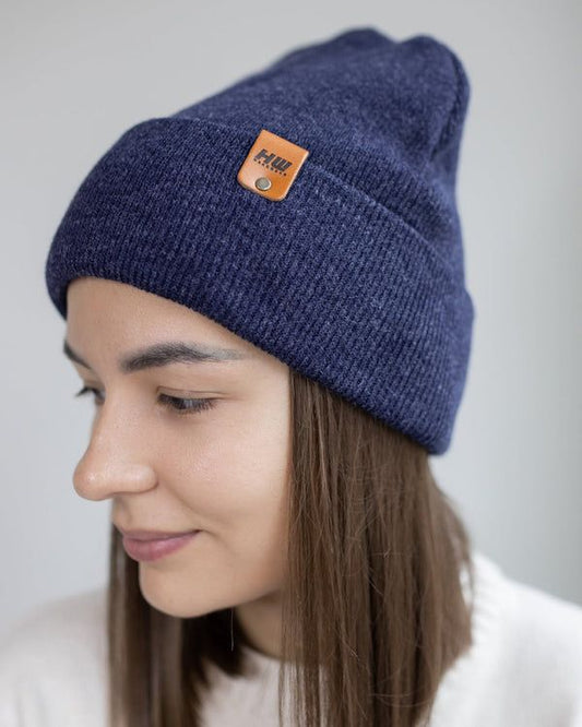 Denim blue cuffed knit beanie with small brown logo patch, worn turned to the side