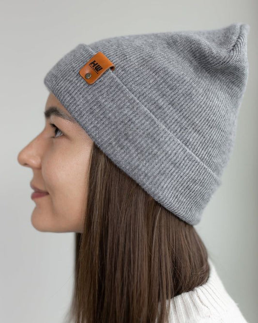 Gray knit beanie on person, folded cuff and orange logo patch detail near side.