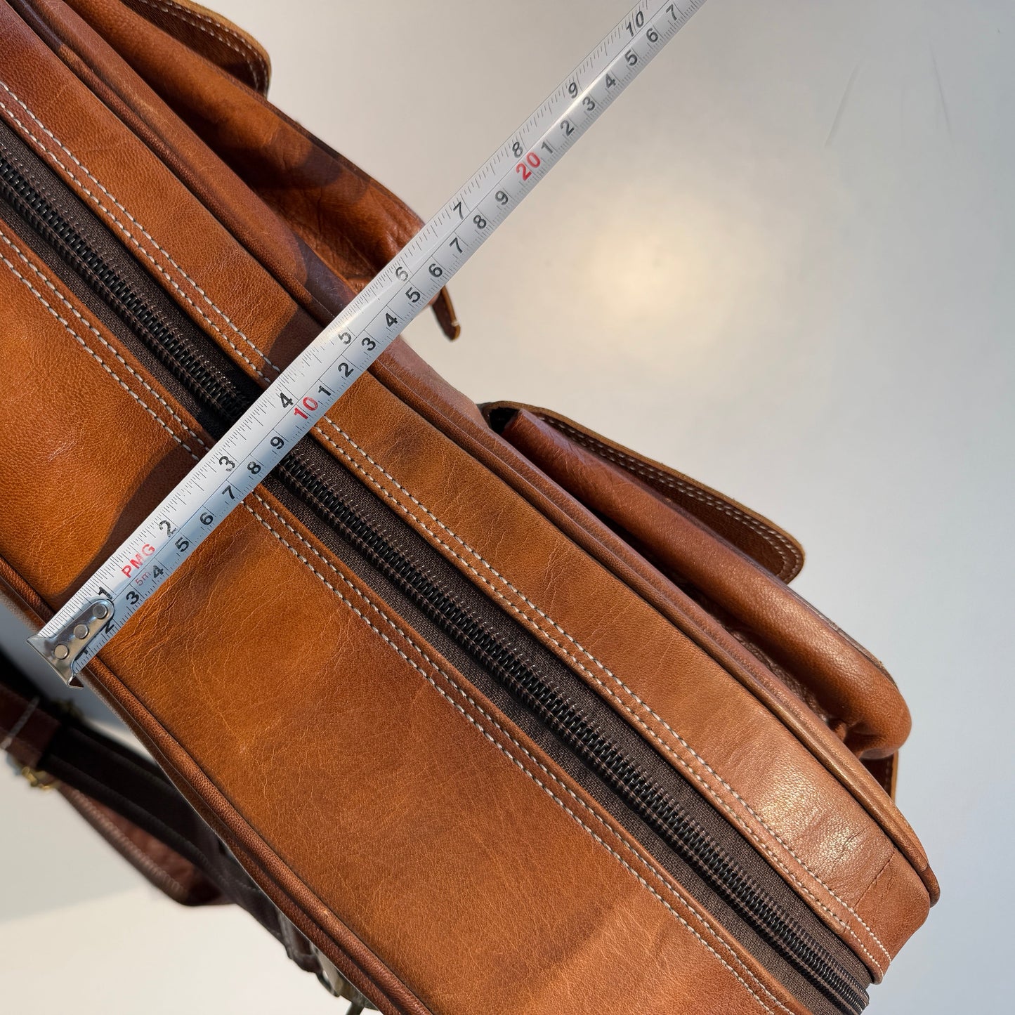 Brown leather guitar case with zippered compartments and stitched panels, measuring tape detail.