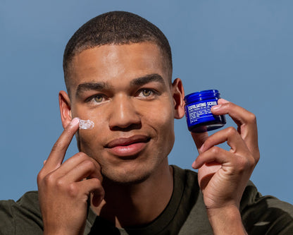 Man applying exfoliating scrub from blue jar to cheek, white scrub visible