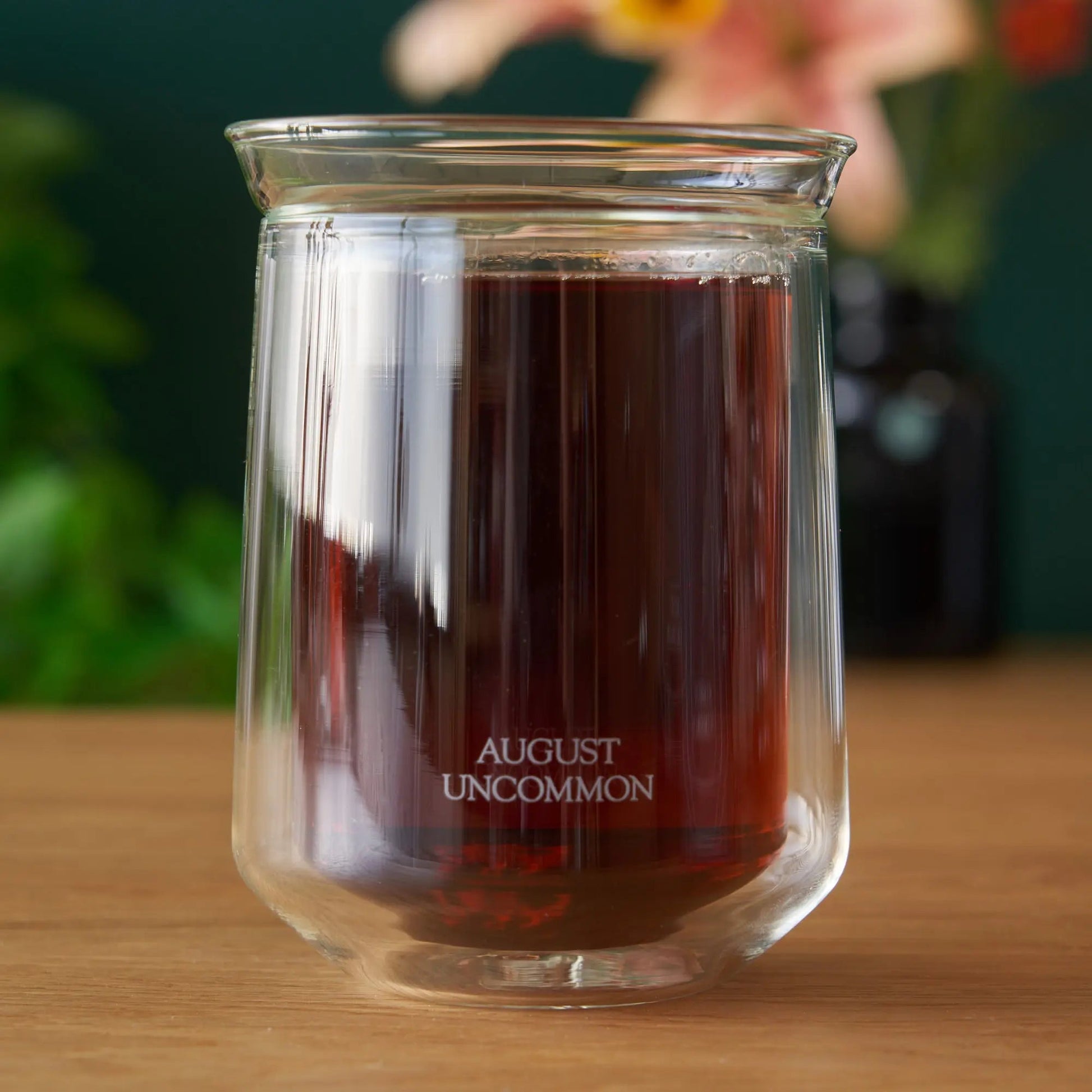 Clear tasting glass with dark brown liquid inside; round rim, AUGUST UNCOMMON etched