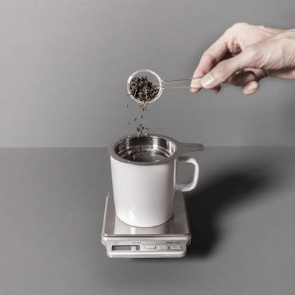 Digital kitchen scale with white mug on top, hand pouring tea leaves into it