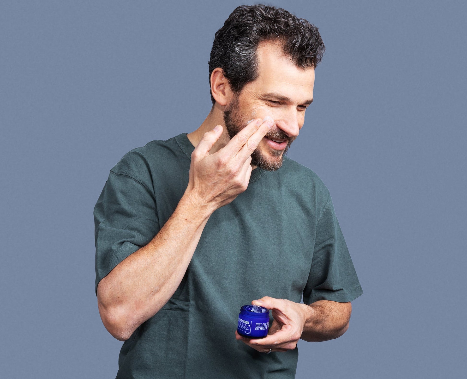 Man rubbing exfoliating scrub on face while holding blue jar