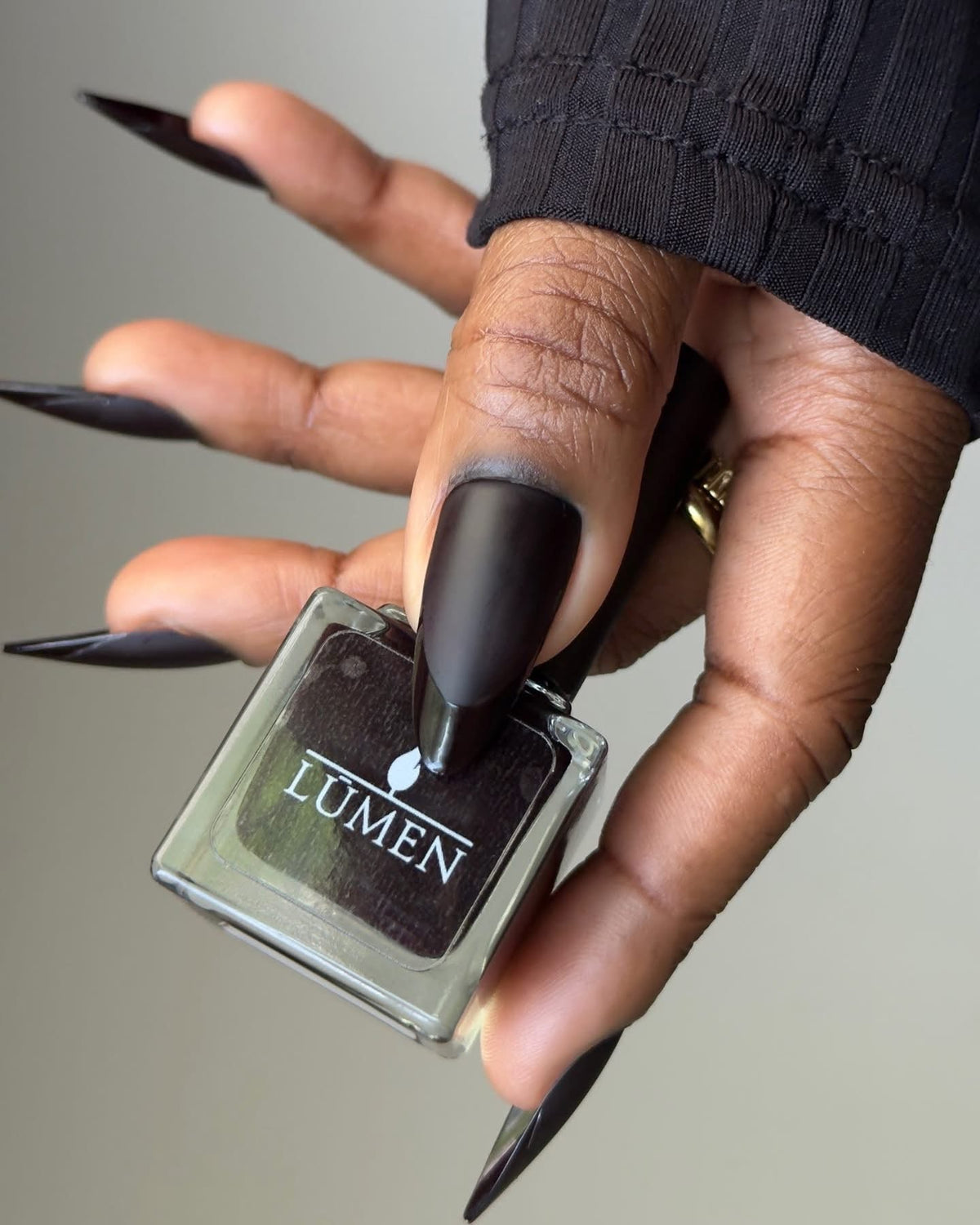 Nail polish bottle with dark shade; long matte nails and ring visible