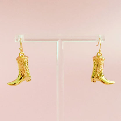 Gold-tone cowgirl boot dangle earrings hanging from a clear display