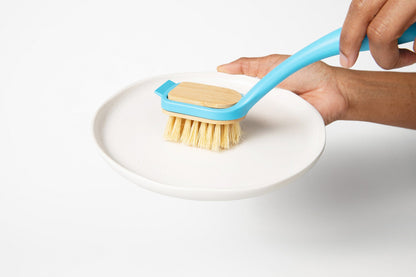 Hand using a blue dish brush over a white dish with light yellow bristles