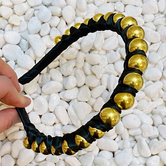 Satin wrapped headband, black with gold round bead accents, laid on white pebbles.
