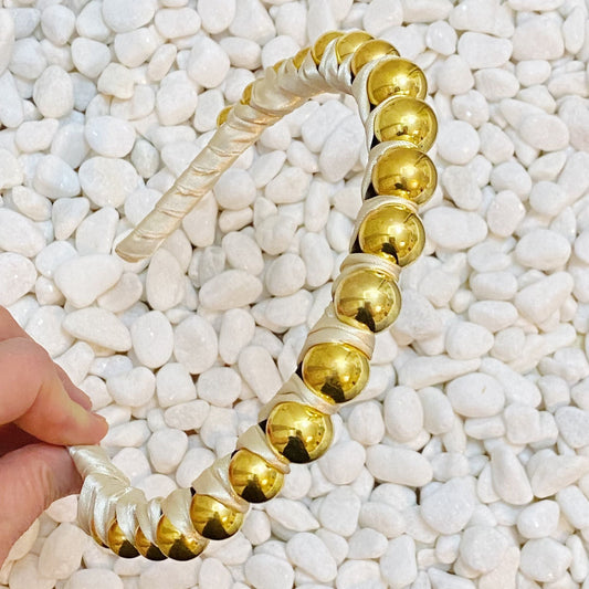 Satin wrapped headband, cream with gold round bead accents, laid on white pebbles.