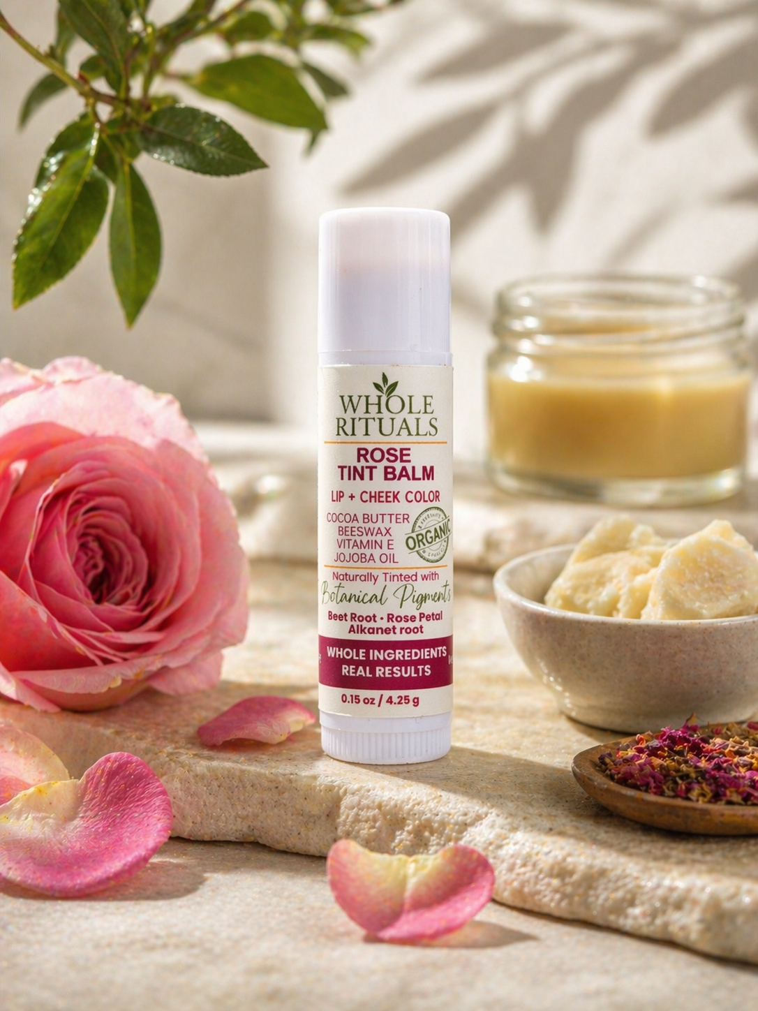 Rose tint balm tube beside pink roses and petals on a stone surface