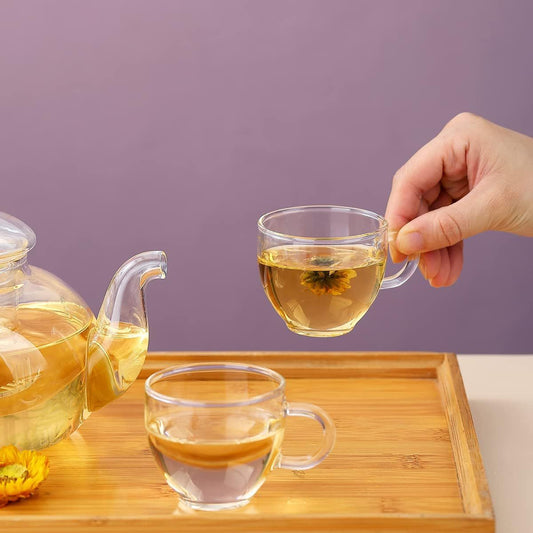Glass tea pot and two tea cups; one cup held above a wooden tray