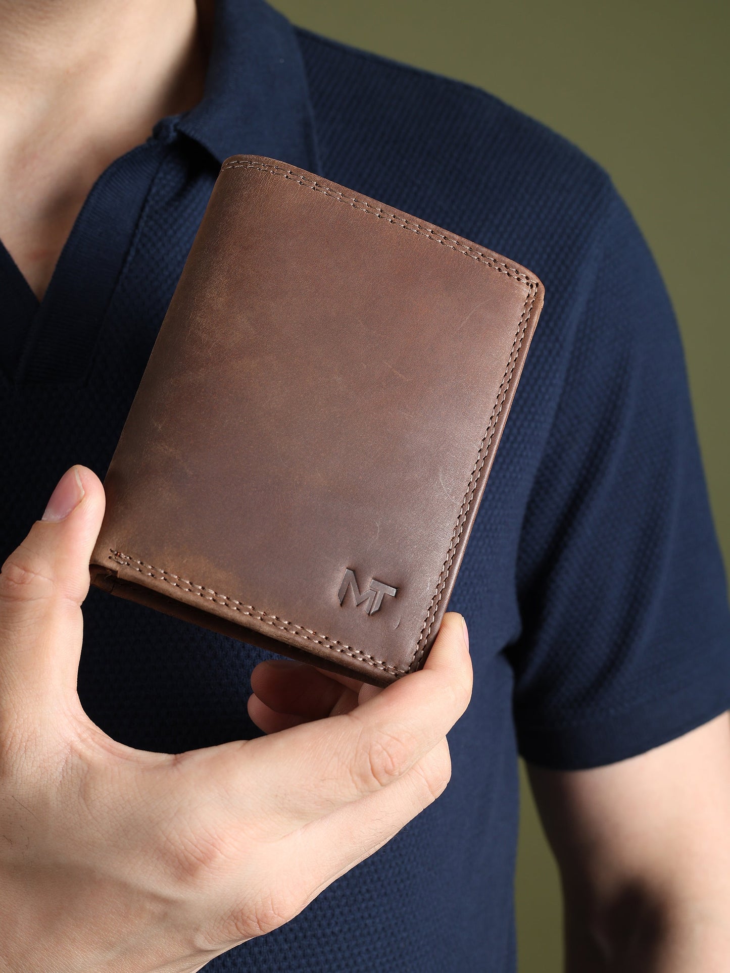 Men's brown leather wallet held in hand, showing stitched front and MT logo.