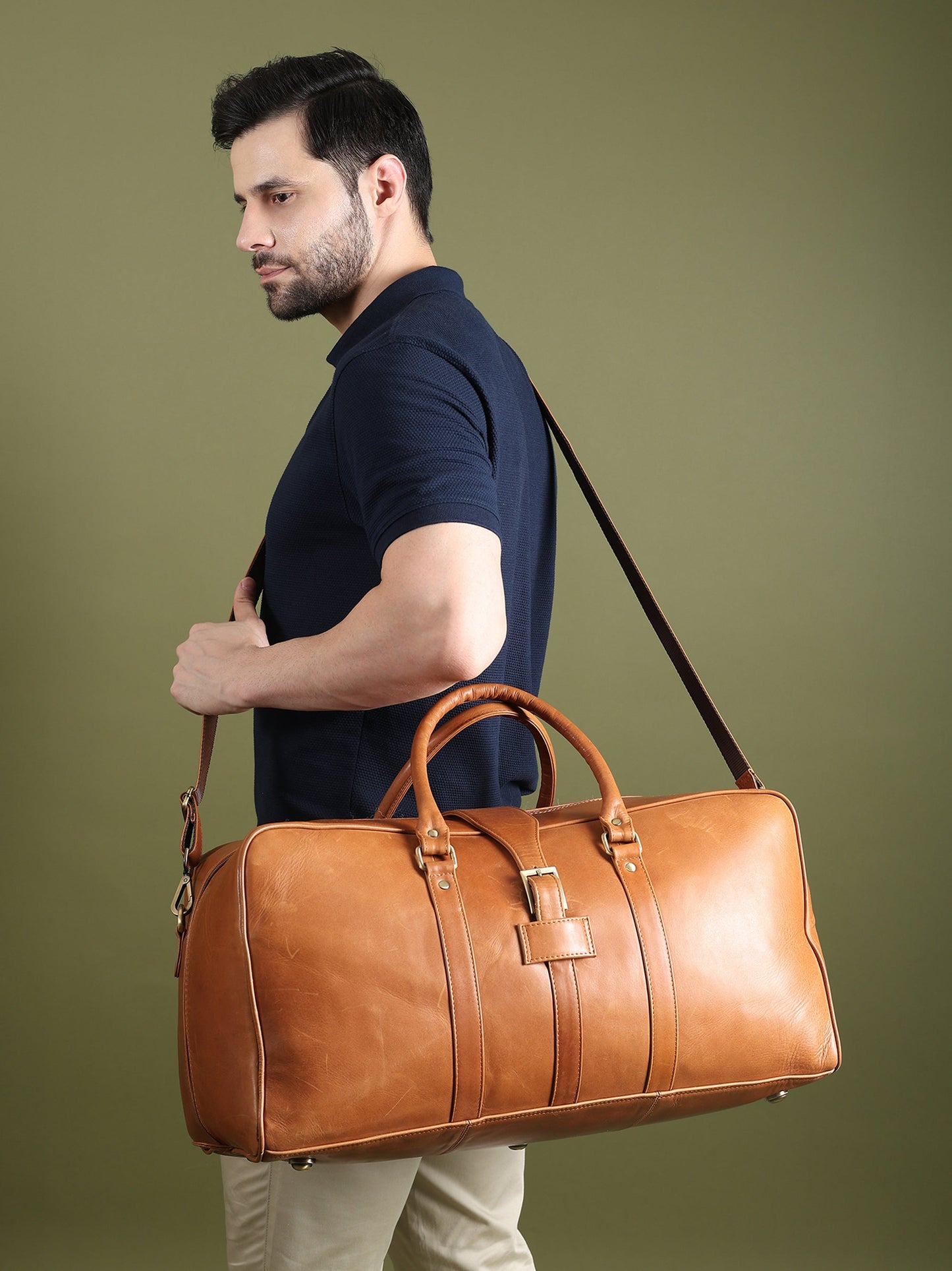 Tan leather duffle bag with long shoulder strap, carried at the side by a model