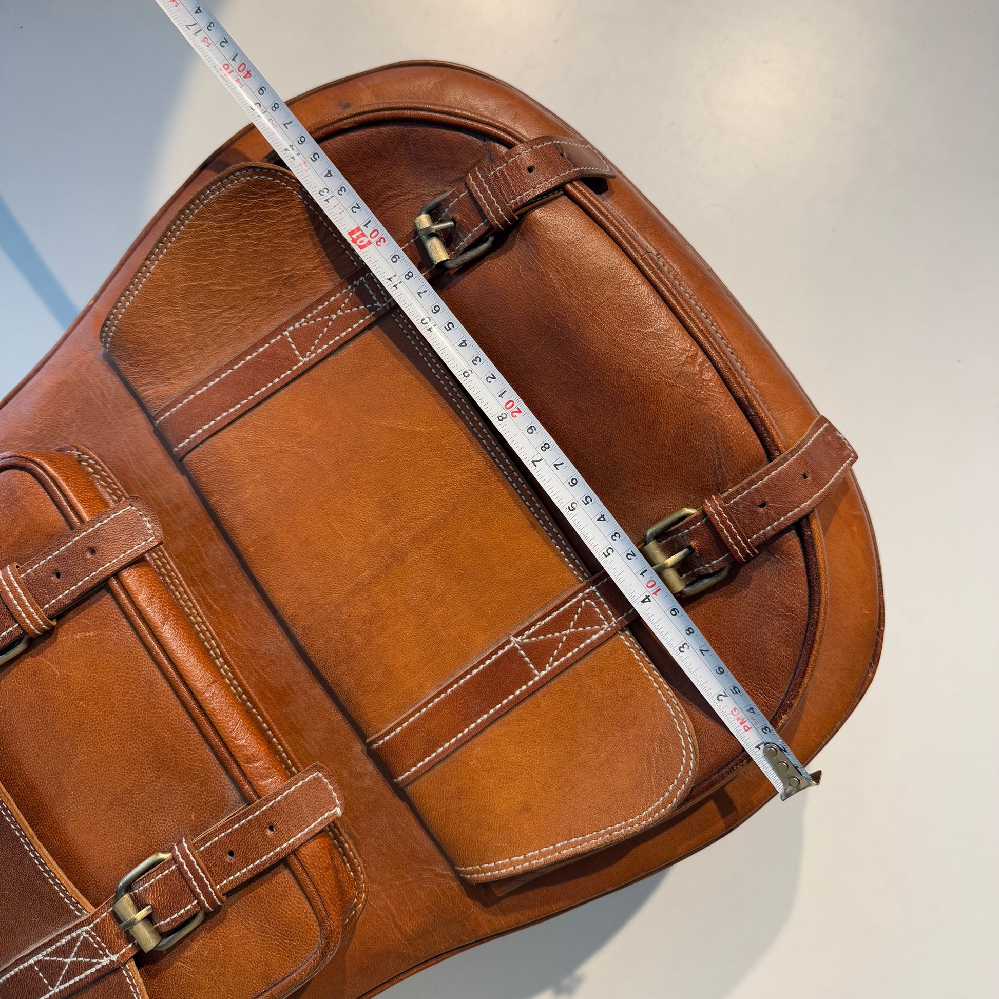 Brown leather guitar case side with handle straps and buckles, measuring tape along body.