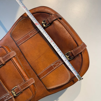 Brown leather guitar case side with handle straps and buckles, measuring tape along body.