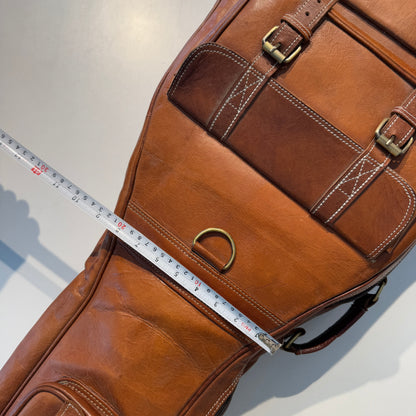Brown leather guitar case close-up with side pocket, buckle strap, and measuring tape.