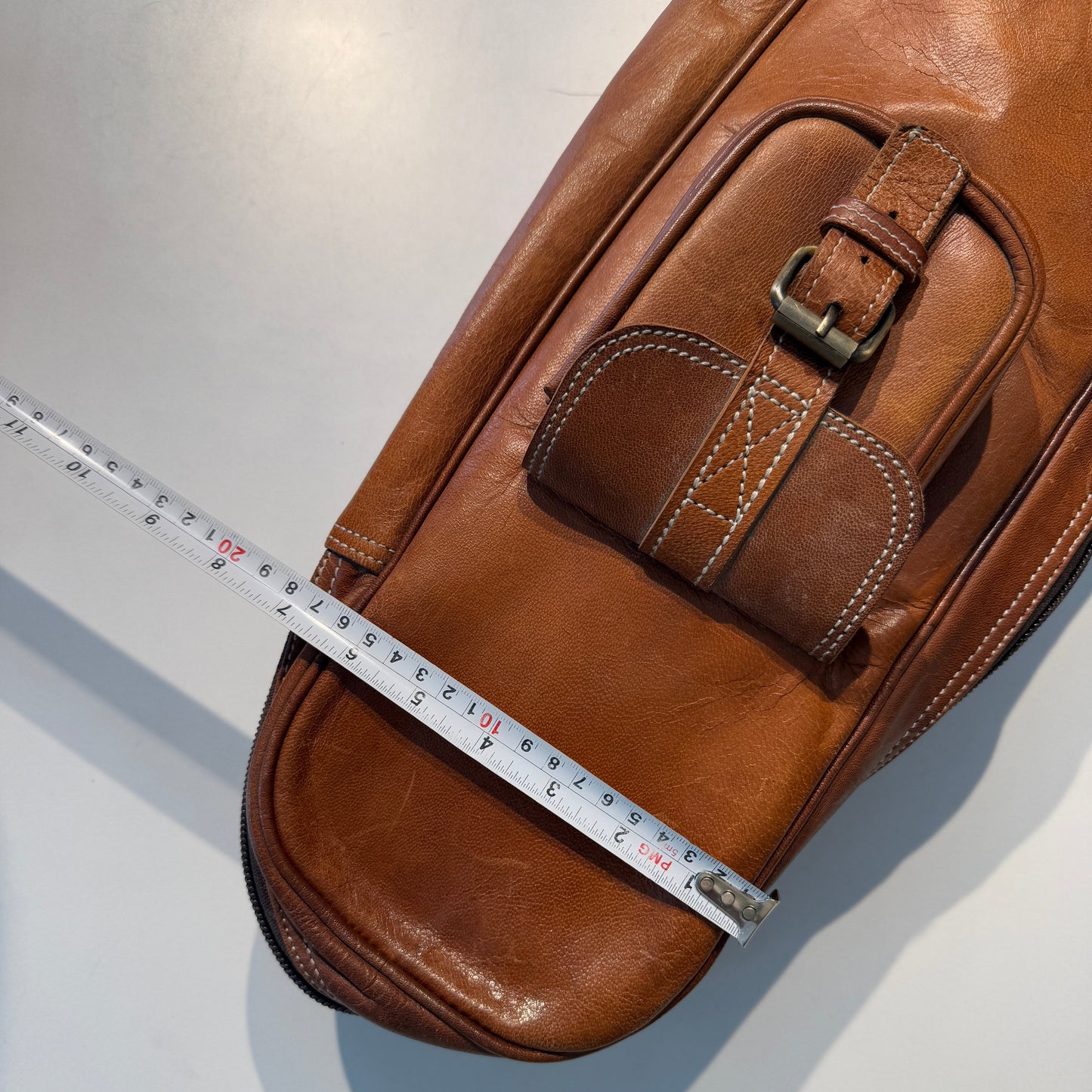 Brown leather guitar case front top pocket with buckle and stitched panel, measuring tape across.