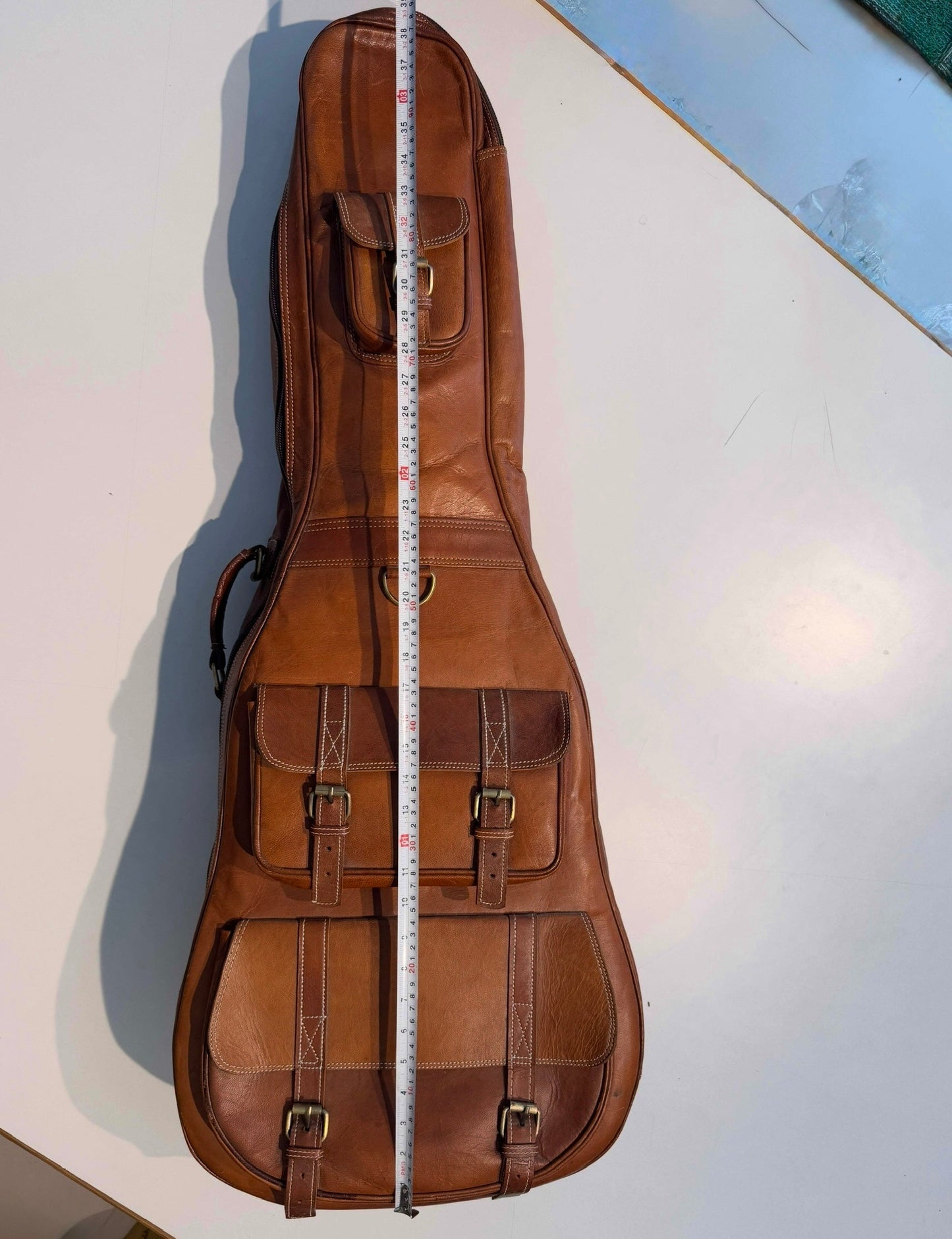 Brown leather guitar case with two front pockets and buckles, measuring tape down the center.