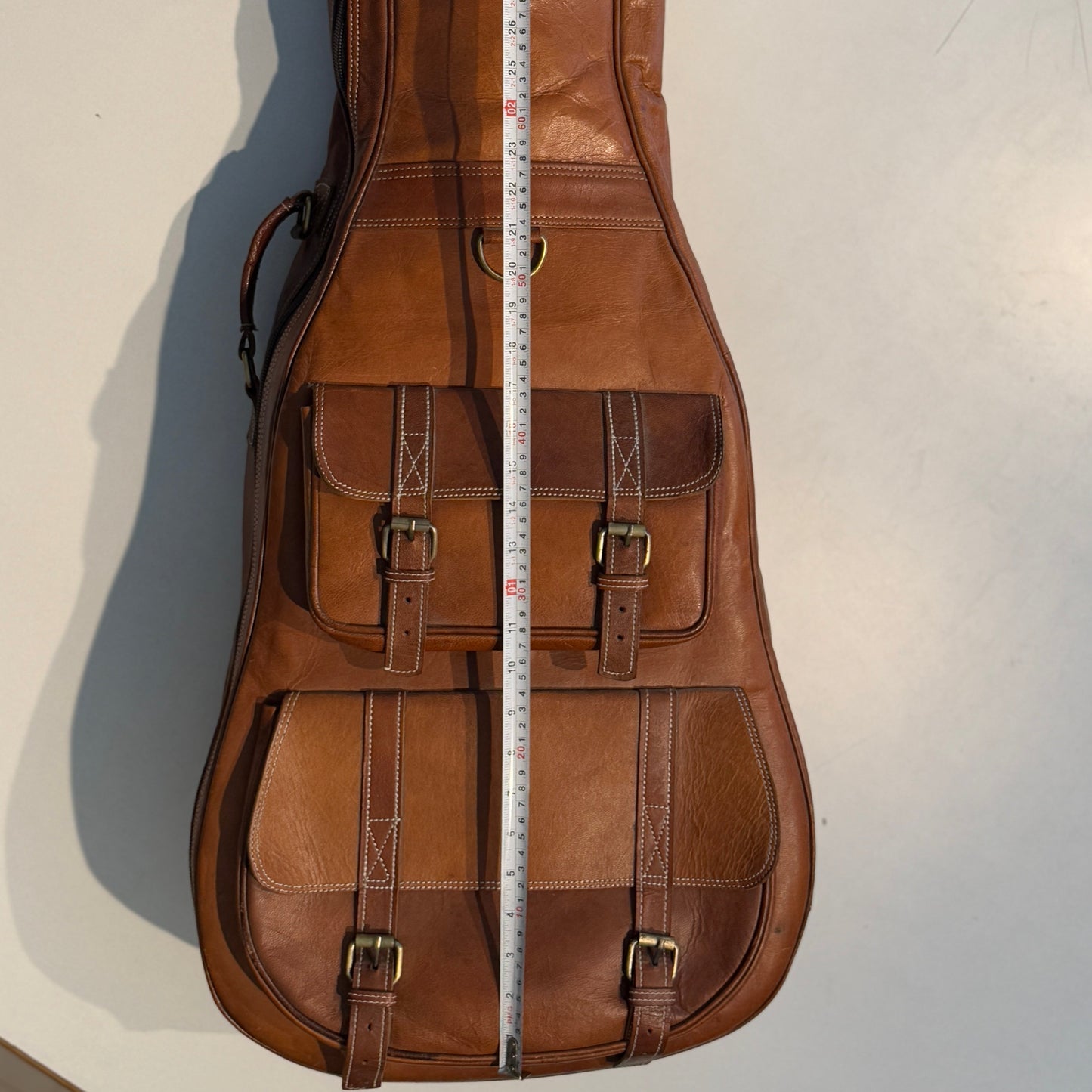 Brown leather guitar case with front buckled pockets and measuring tape at center.