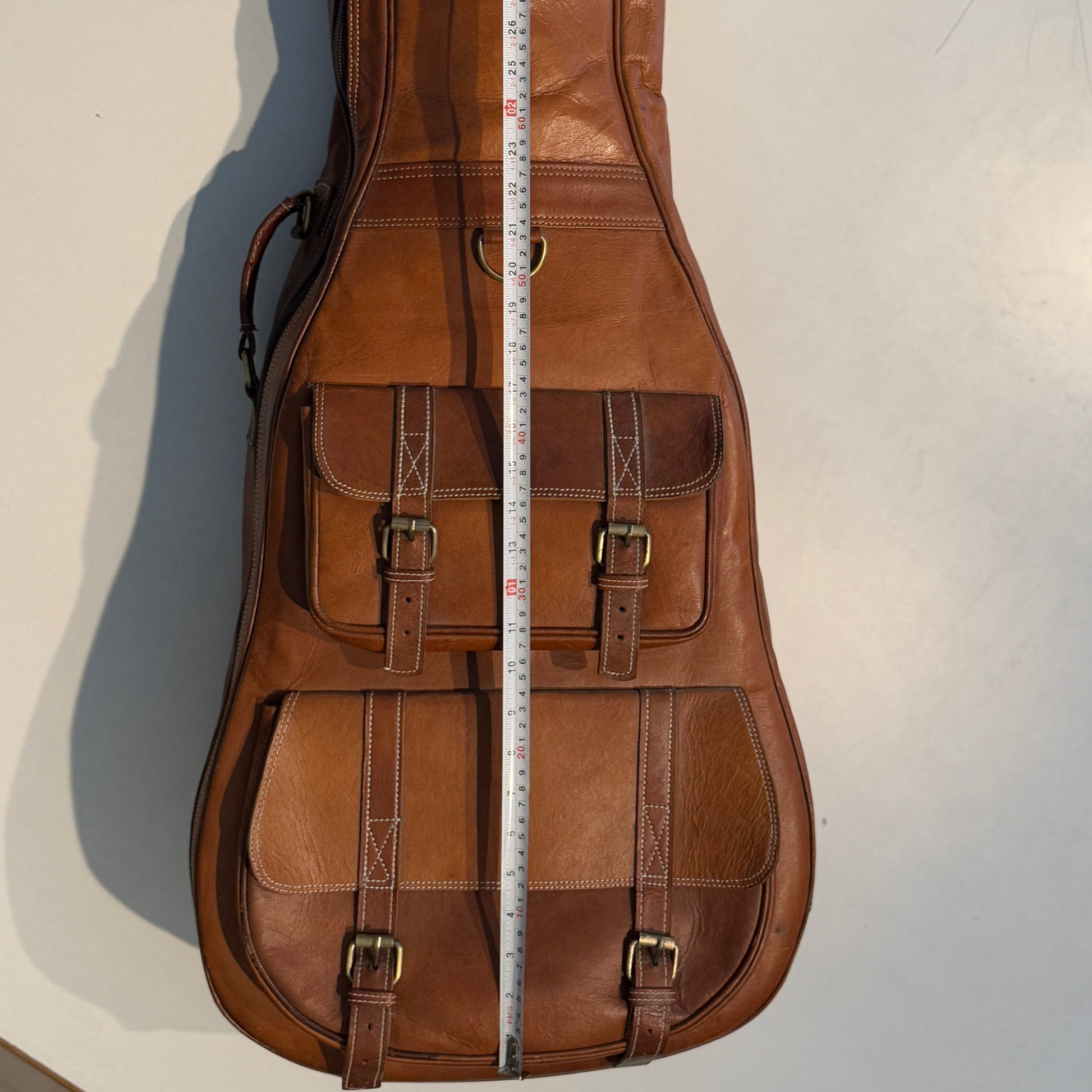 Brown leather guitar case with front buckled pockets and measuring tape at center.