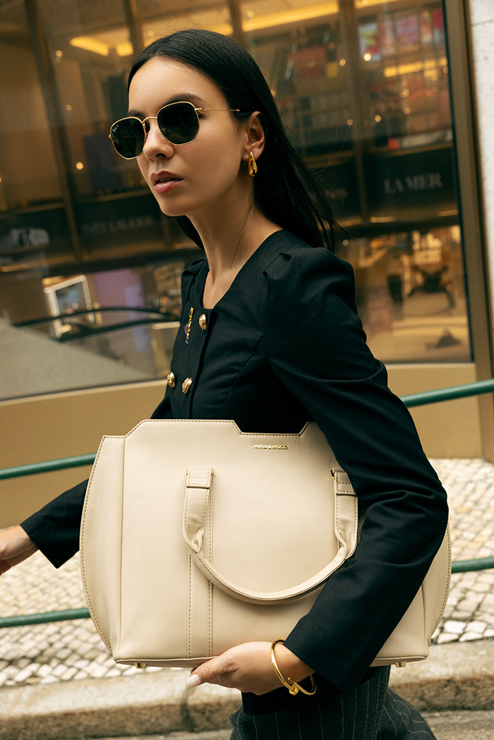 Light taupe crossbody bag carried by a model with round shoulder strap and gold clasp