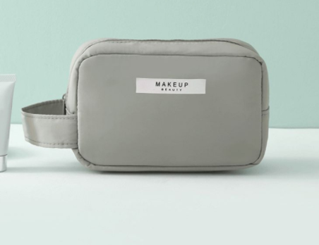Cosmetic pouch light gray, zip-top with white front label and rounded rectangular shape.