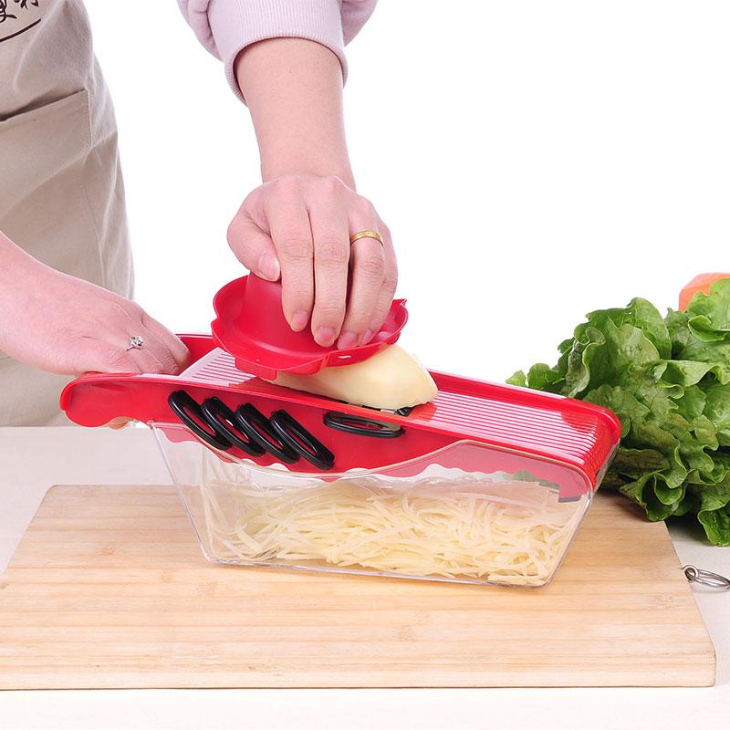 Red kitchen slicer with protective guard and blade plate, shown cutting potatoes on tray.