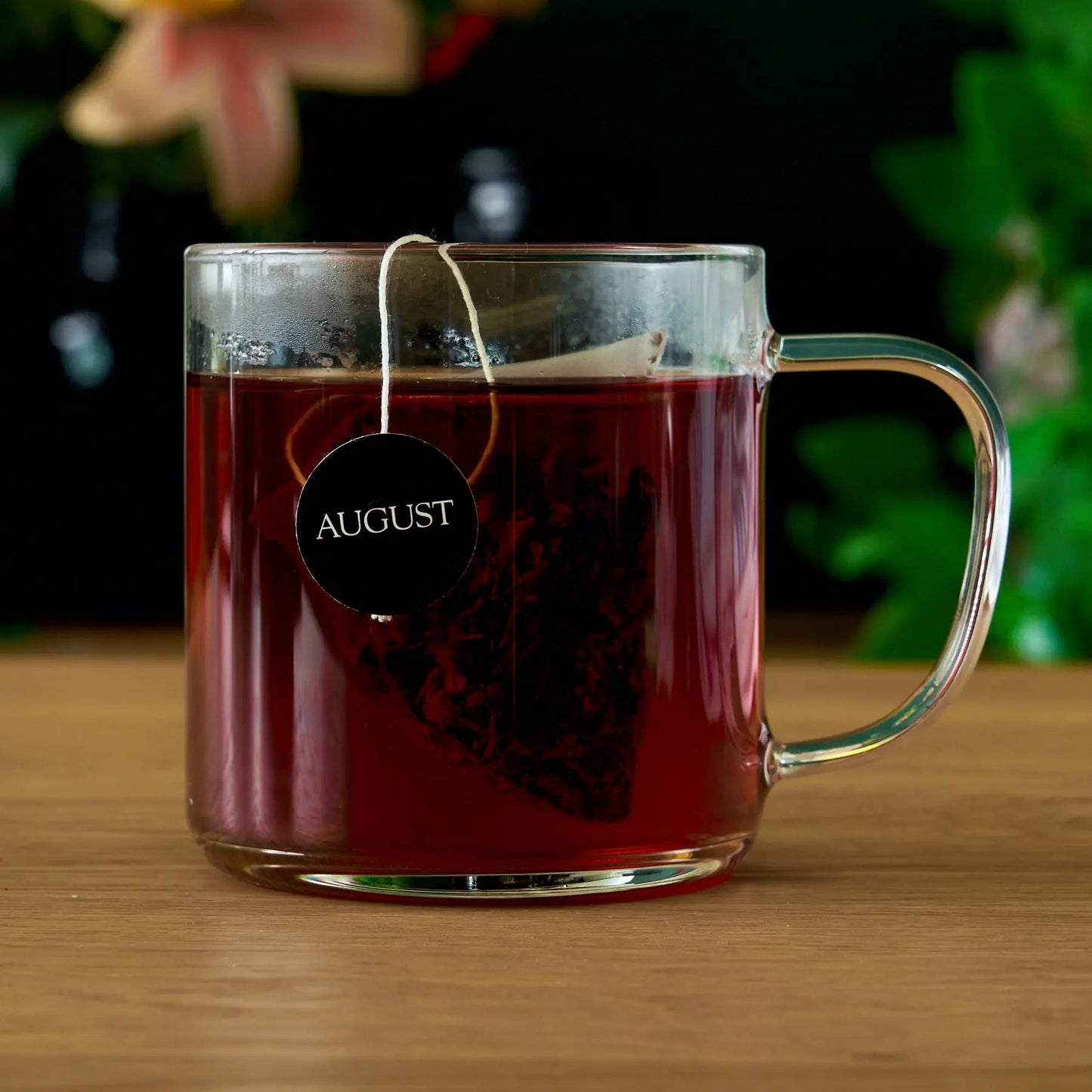 Glass mug of dark brewed tea with a pyramid tea bag tag labeled AUGUST