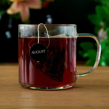 Glass mug of dark brewed tea with a pyramid tea bag tag labeled AUGUST