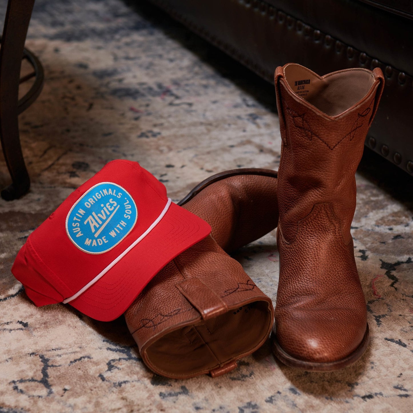 Red snapback hat with blue oval logo patch, shown with brown boots on a floor.