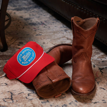 Red snapback hat with blue oval logo patch, shown with brown boots on a floor.