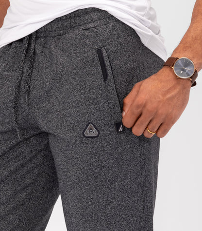 Dark charcoal gray athletic sweatpants close-up: elastic waistband, logo patch, side pocket seam.