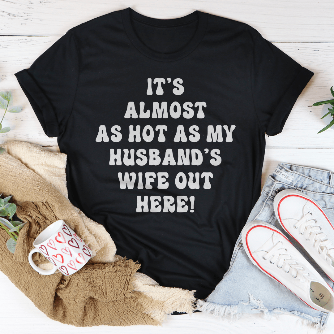 Black graphic T-shirt with 'It's almost as hot as my husband's wife out here!'