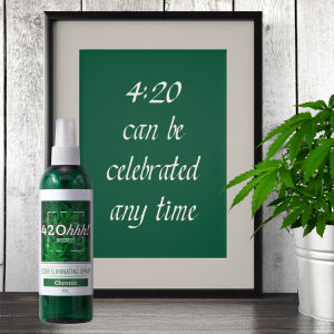 Green spray bottle odor remover with white cap on a tabletop beside a framed sign and plant.