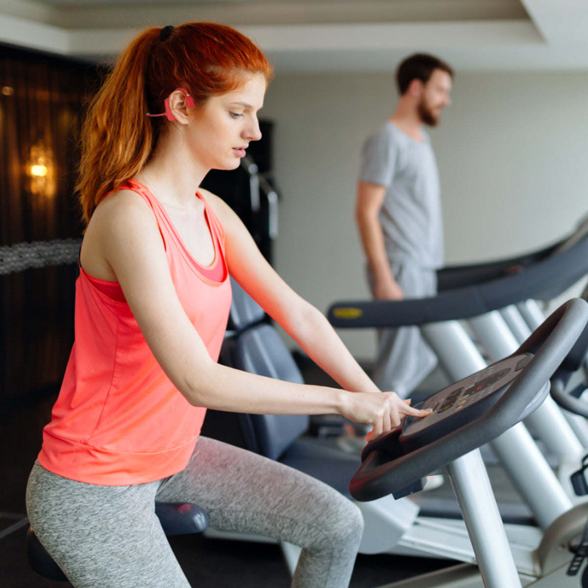 Red open-ear wireless headphones worn during treadmill workout, earbud module visible.