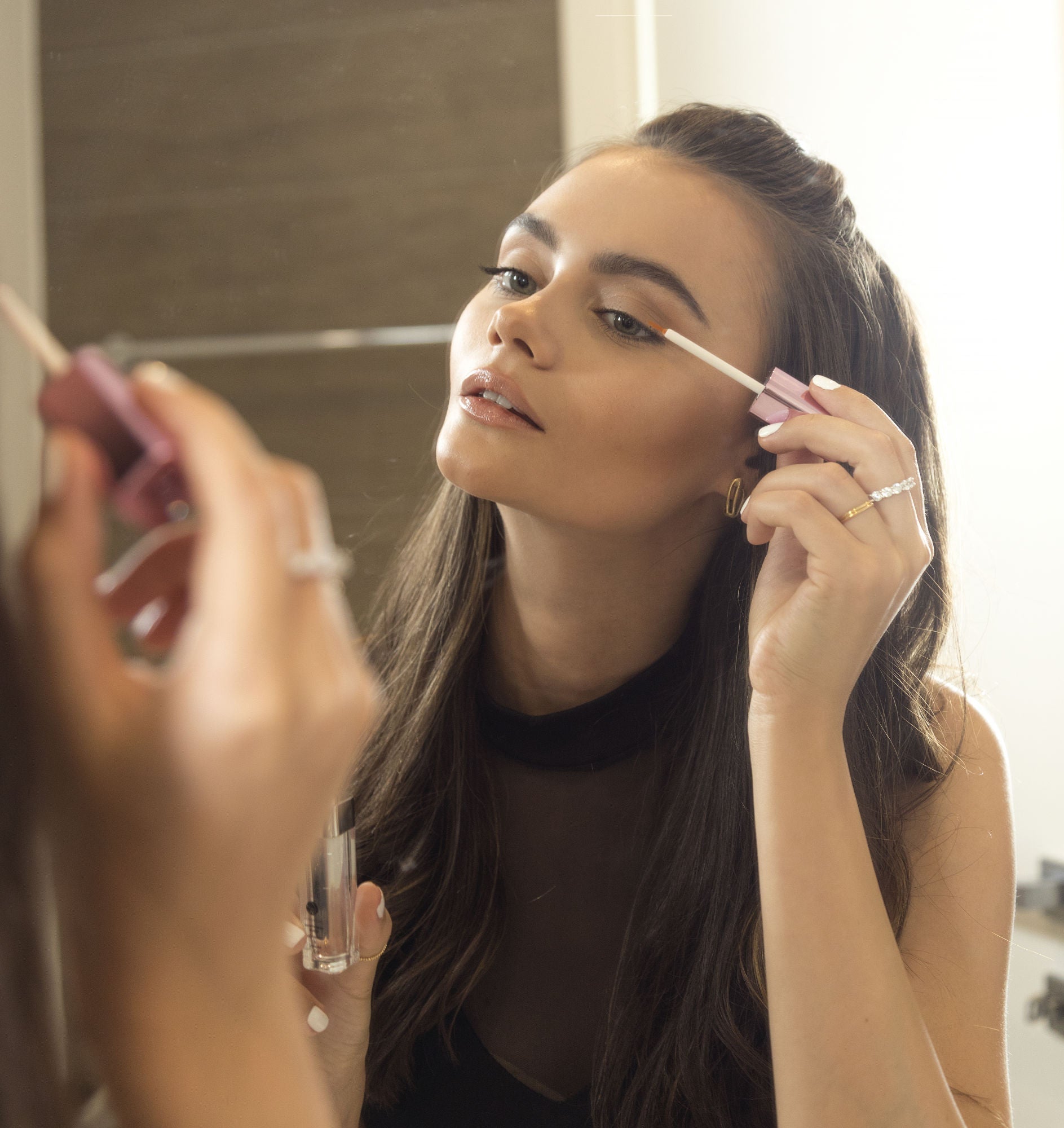 Woman applying eyelash serum with applicator brush near her eye; eyelash serum bottle visible.
