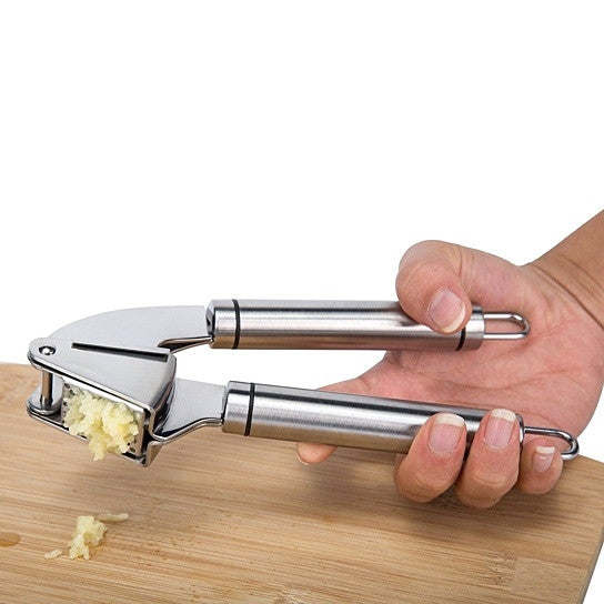 Hand using a stainless steel garlic press on a wooden board with garlic pieces.