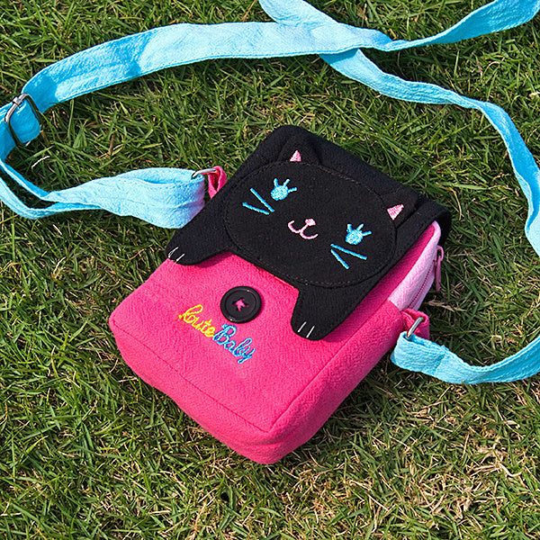 Embroidered cat swingpack bag with pink body, black flap, and light blue shoulder strap.
