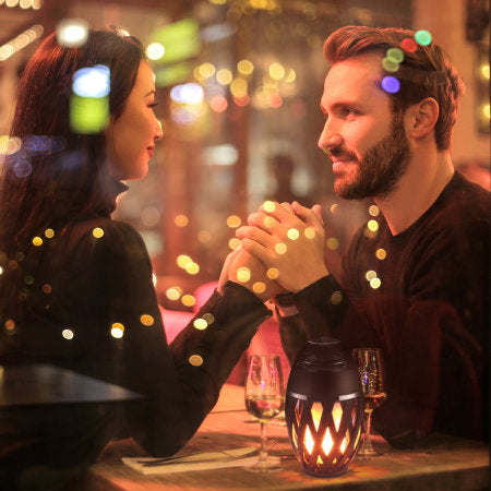Outdoor LED torch speaker with glowing amber flame-pattern, black lantern on tabletop.