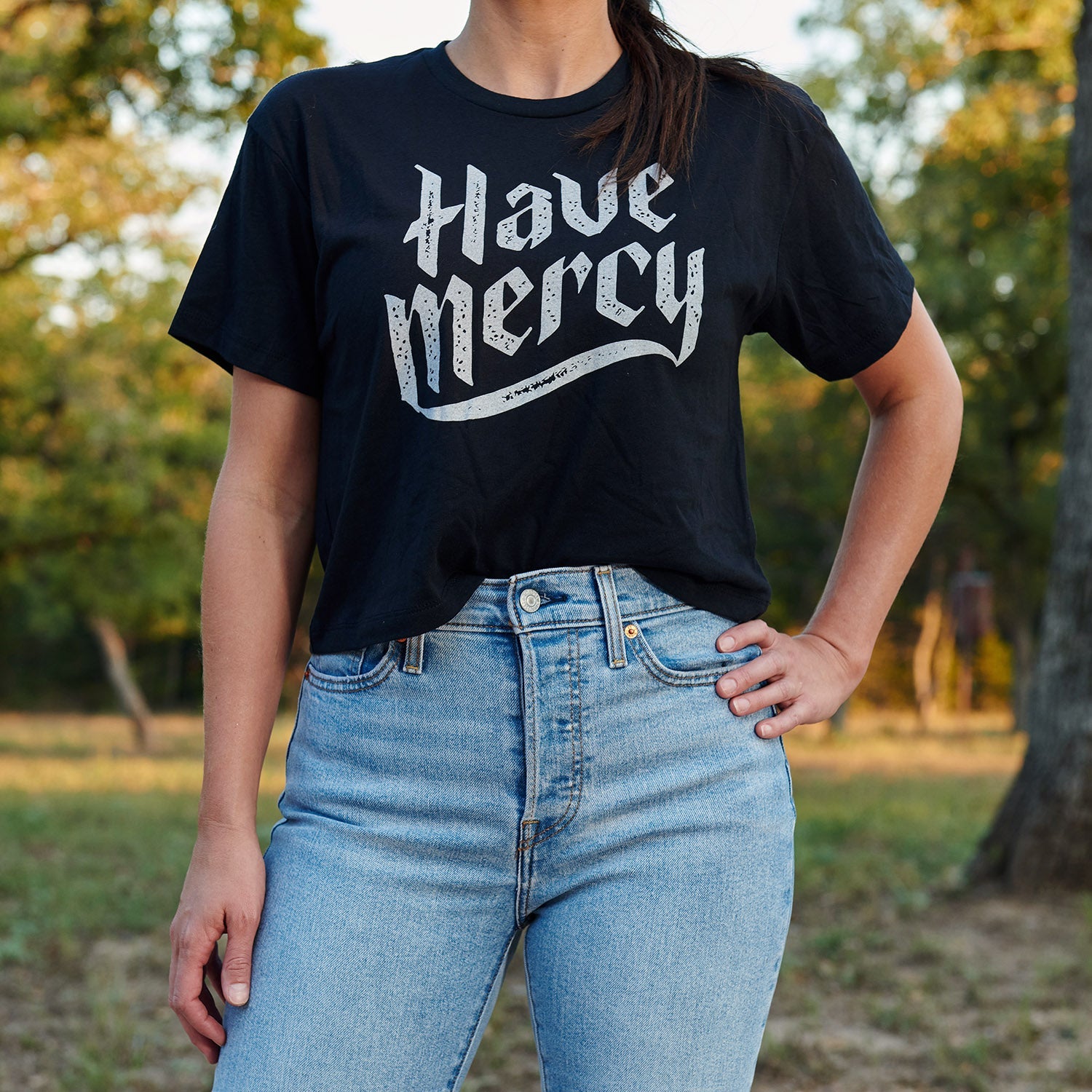 Dark cropped T-shirt with short sleeves, "Have mercy" graphic worn with jeans
