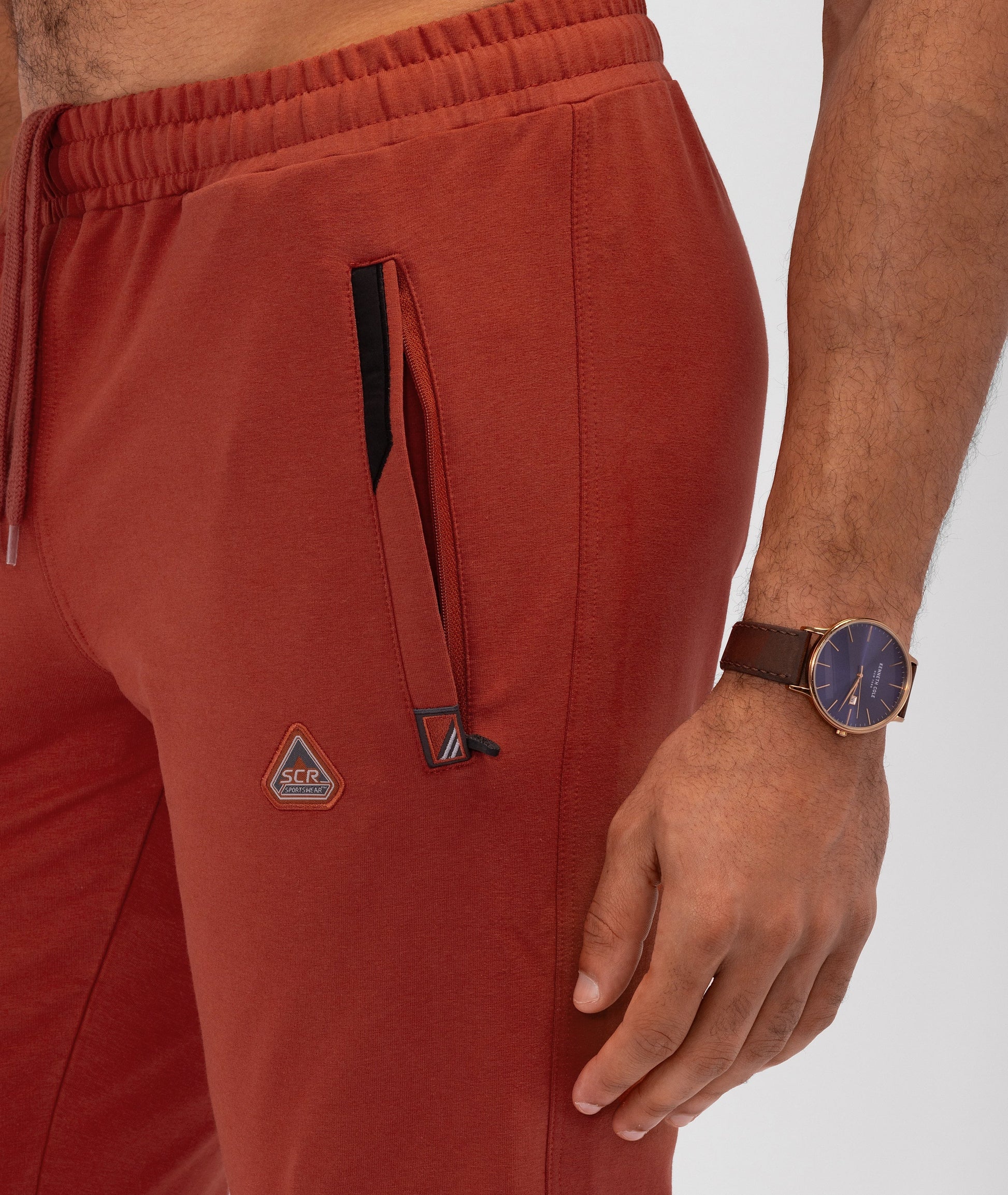 Rust red athletic sweatpants close-up: elastic waistband, drawstring, side zip pocket, logo patch.