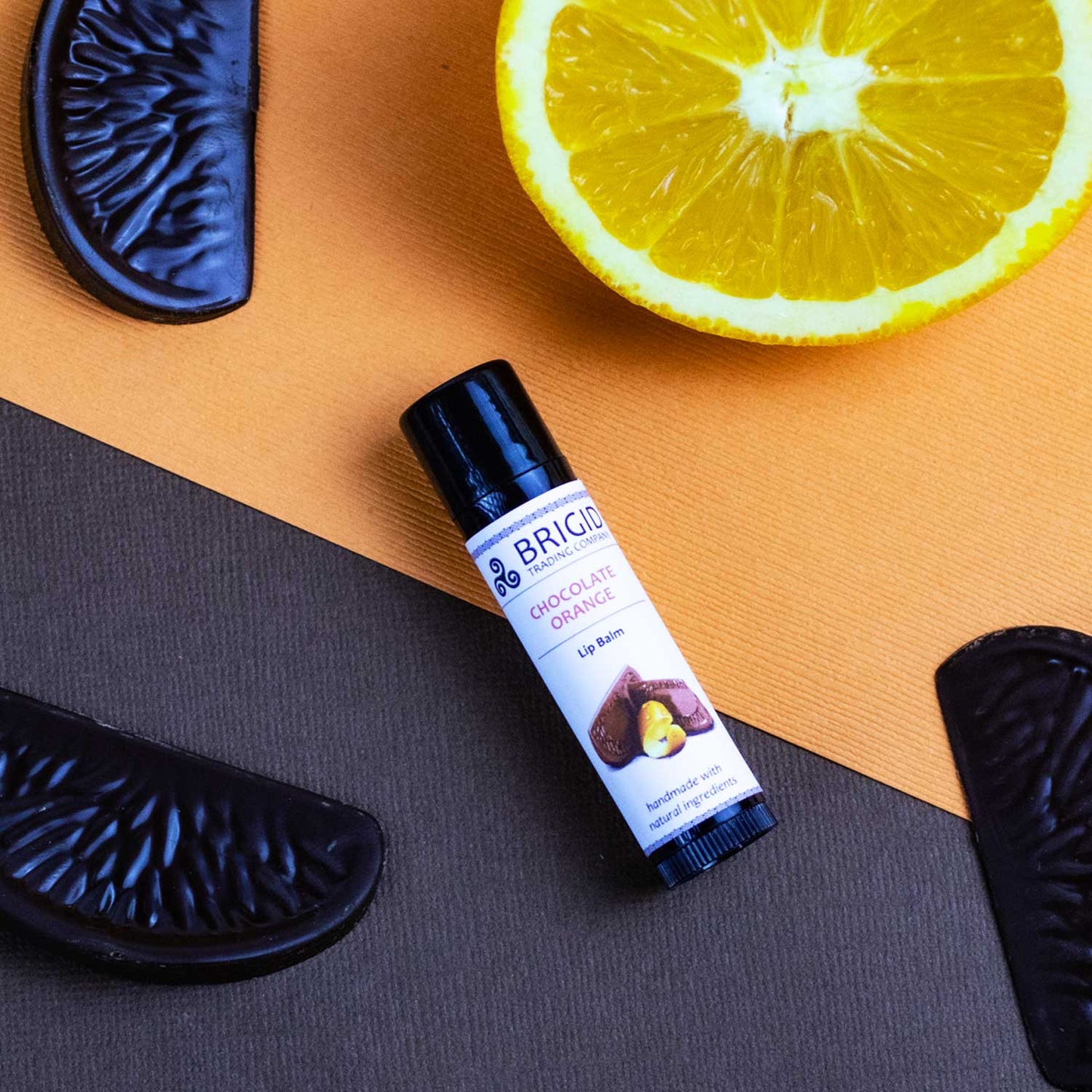 Natural lip balm tube with black cap on tan surface, beside orange slice and dark chocolate.