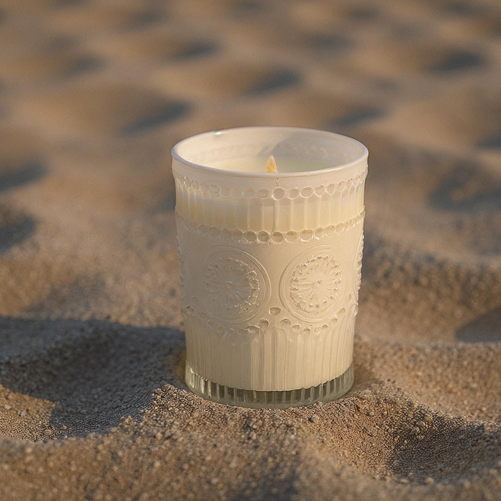 Ivory embossed glass scented candle, lit, on sandy surface
