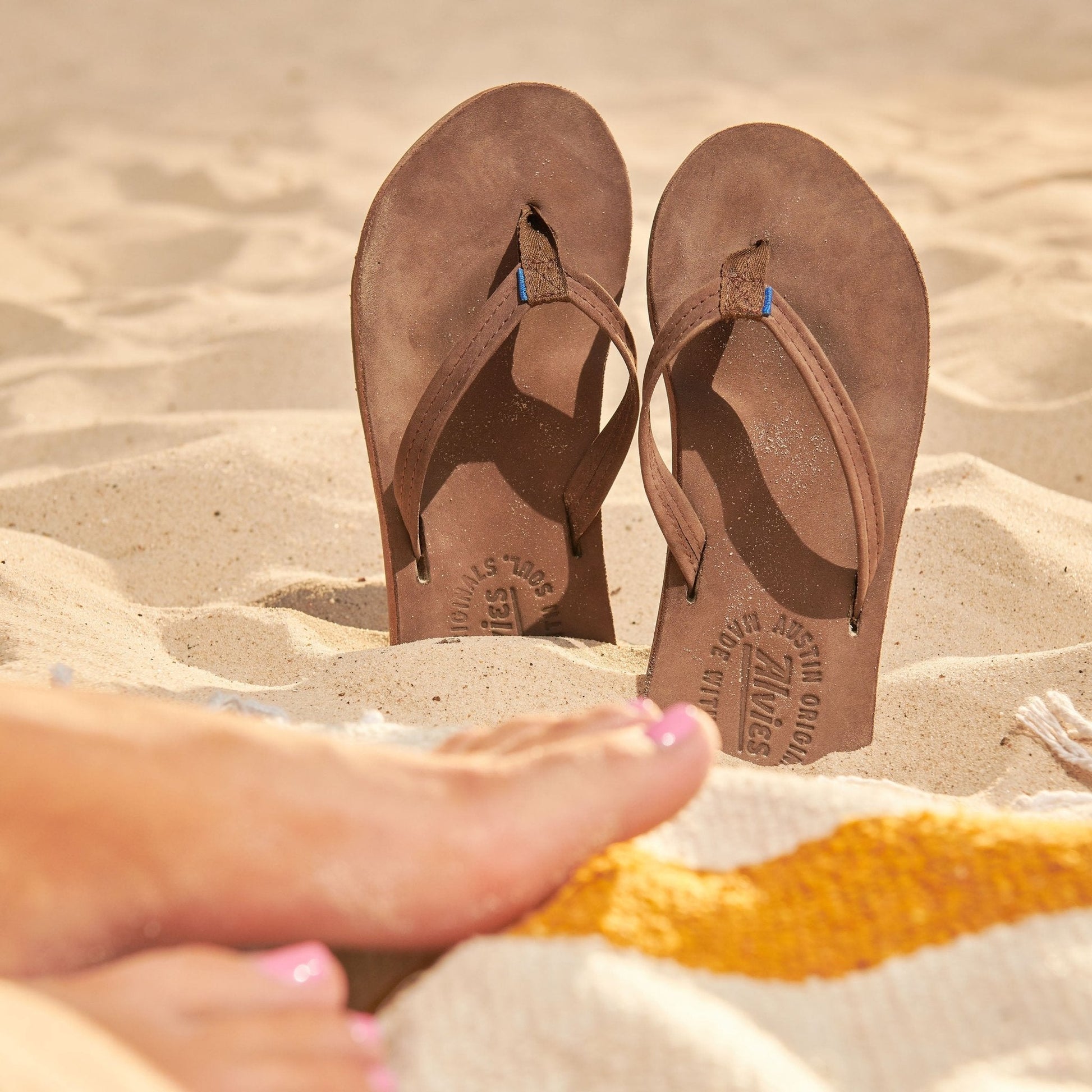 Brown suede flip-flops with toe thongs and blue-accented straps on sand