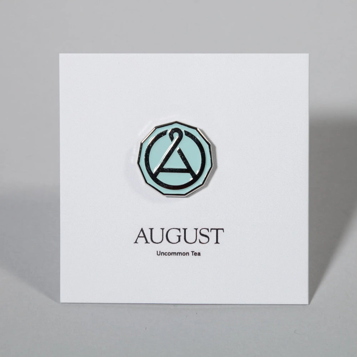 Enamel seal pin with teal octagon border and A emblem on a white card
