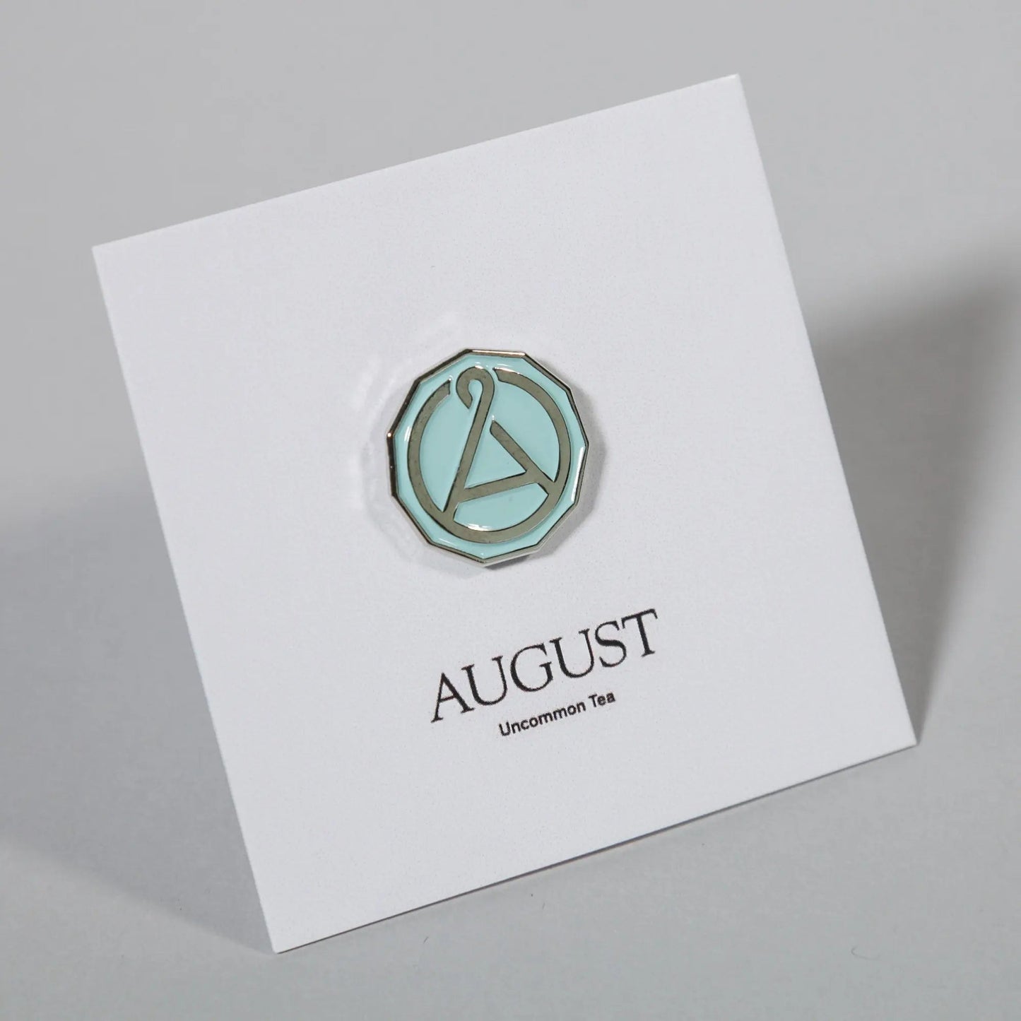 Enamel seal pin with teal octagon border and A emblem on an angled white card