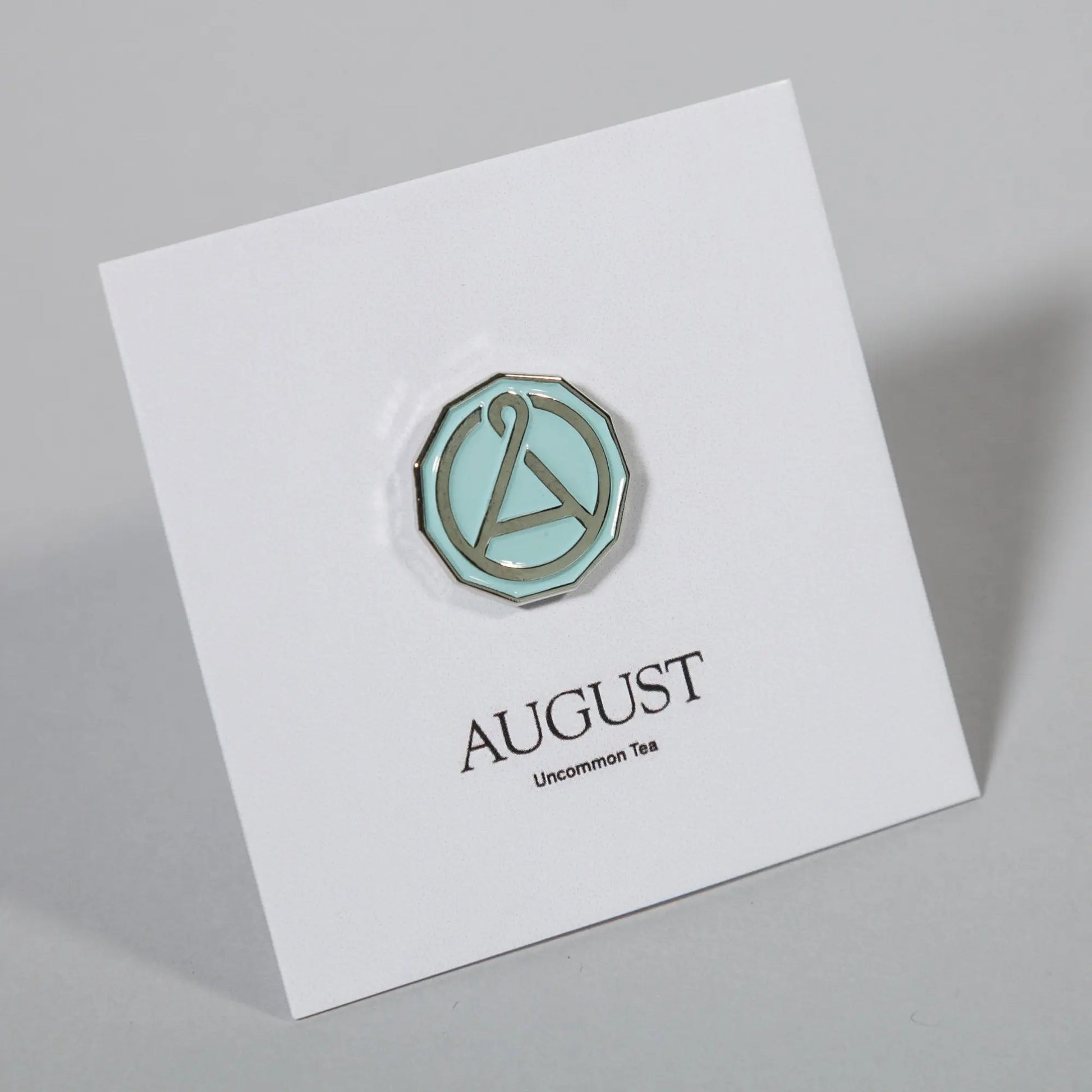 Enamel seal pin with teal octagon border and A emblem on an angled white card