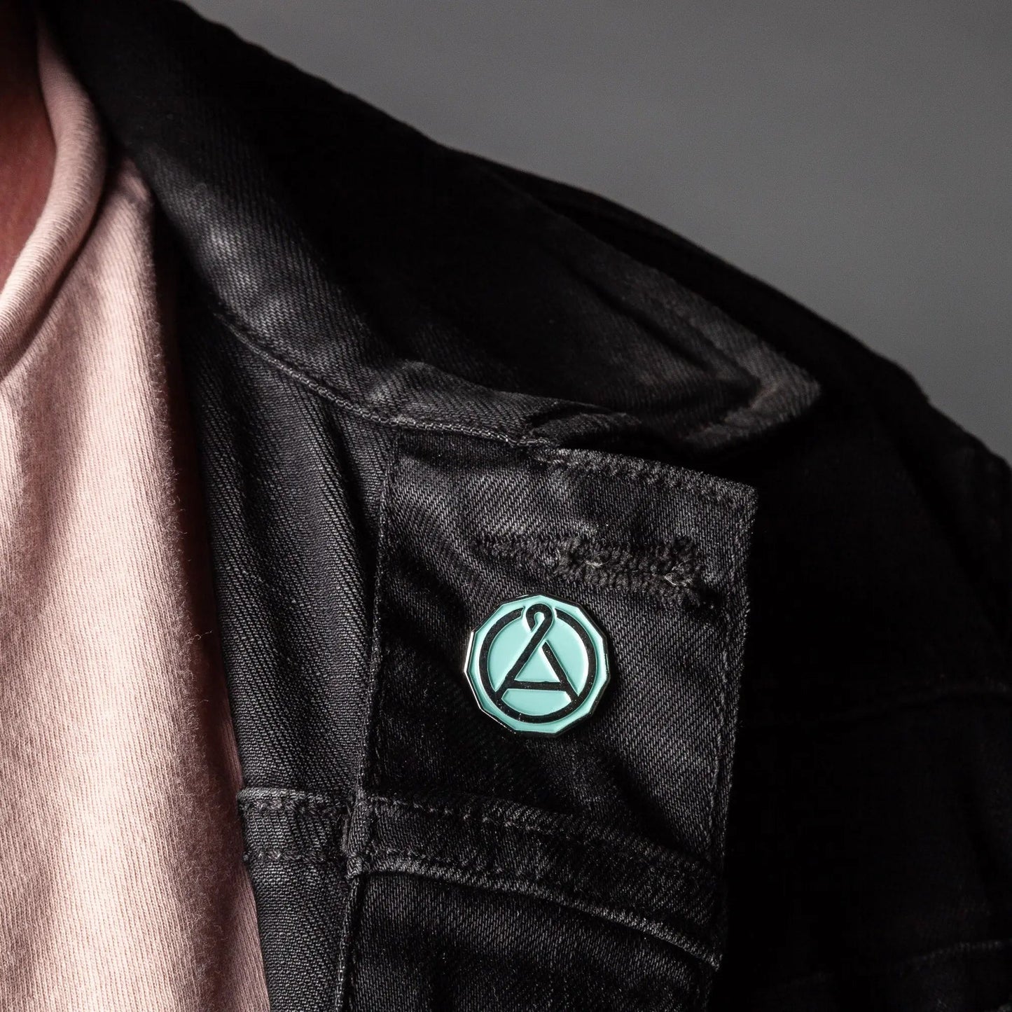 Enamel seal pin with teal octagon outline and A emblem on a dark jacket pocket