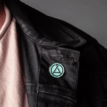 Enamel seal pin with teal octagon outline and A emblem on a dark jacket pocket
