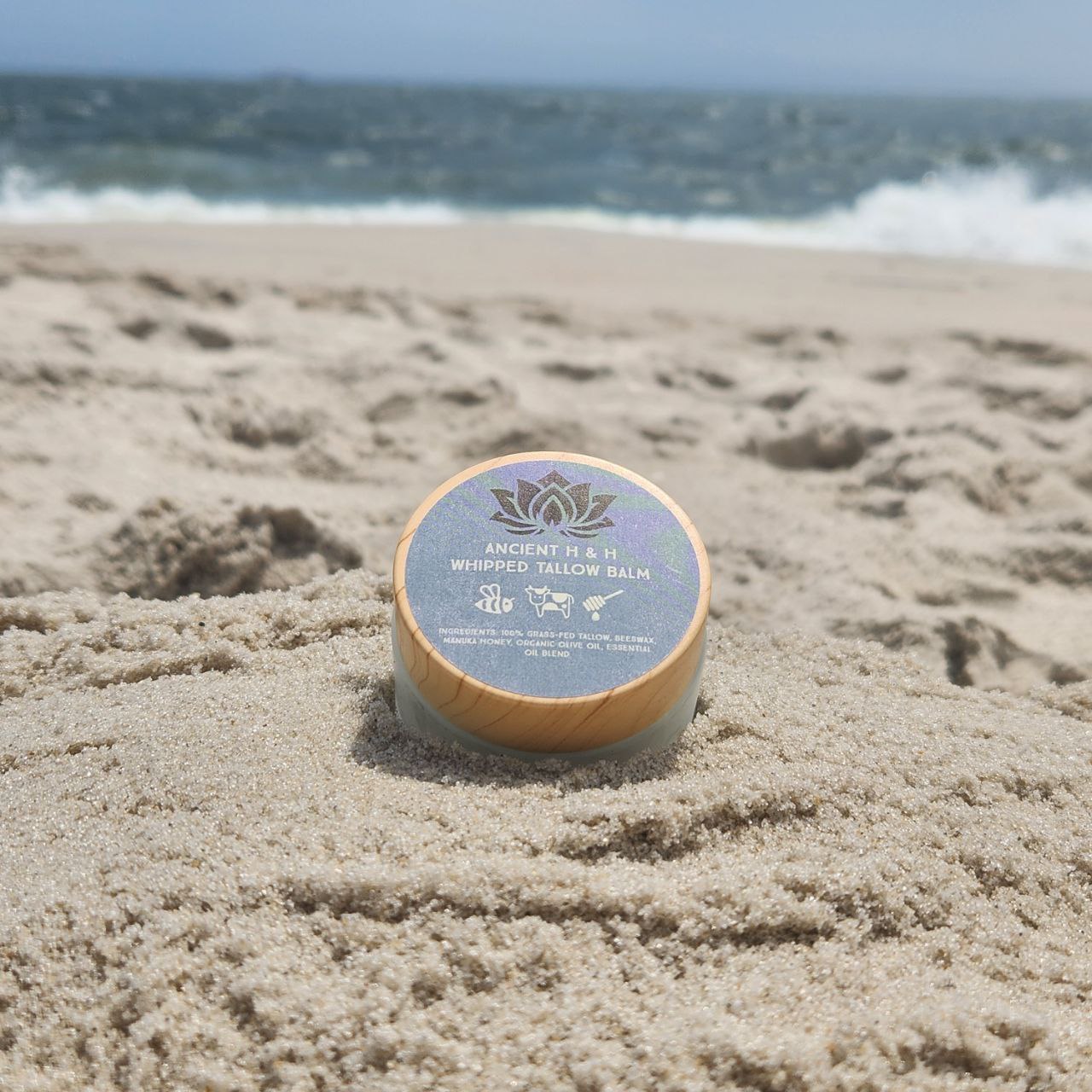 Tallow balm jar on sandy surface with blue lid label and gold lotus emblem
